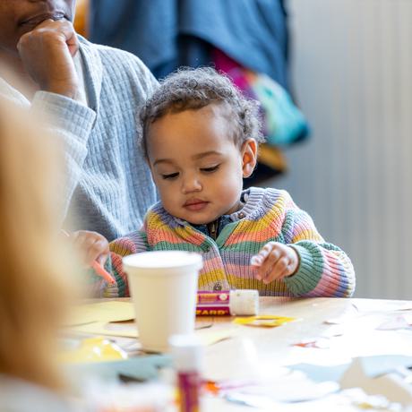 Baby doing crafting at a table sat on the knee of their parent/carer