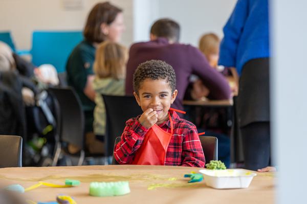 Young boy in checked red top crafting at a table, hand is next to mouth and he is smiling