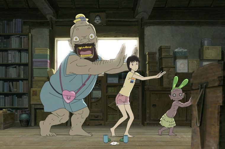 A scene from the animated film 'A Letter to Momo' showing three characters in a cluttered room filled with bookshelves and stacked boxes. On the left is a large, muscular spirit with a yellow hat, blue shirt, and pink bag, gesturing with outstretched arms. In the centre is a young girl with black hair wearing a yellow shirt and pink shorts, also with her arms spread wide. On the right is a smaller spirit character with green rabbit-like ears wearing a yellow skirt. Two blue mugs sit on the wooden floor between them. The room appears to be an attic or storage space with books lining the walls and various items stored throughout.