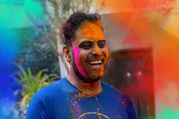 An Indian man in his thirties/forties wearing a blue T-shirt has a large smile on his face. His face is daubed with bright pink and orange paint.