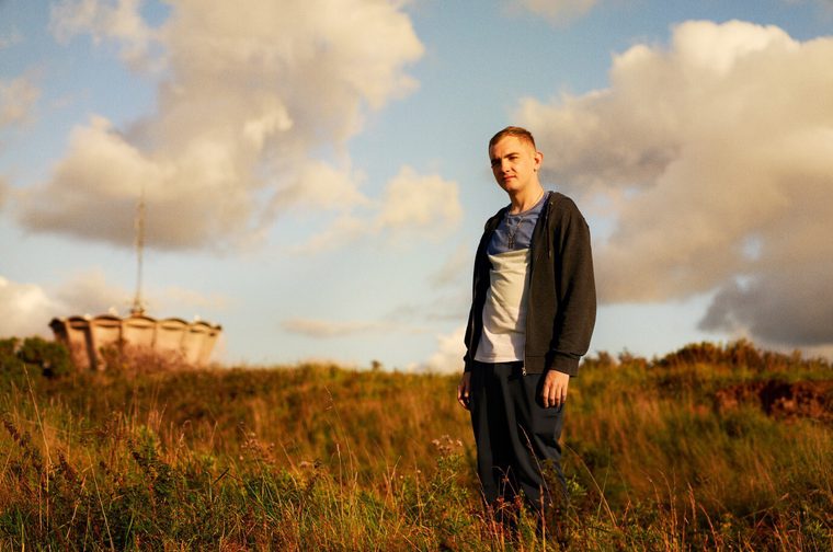 The image shows a young man standing in a field of tall grass and wildflowers, with warm, soft lighting from what appears to be late afternoon or early evening sun. He's wearing a dark zip-up jacket over a light-colored shirt and dark pants, creating a nice contrast against the natural setting.