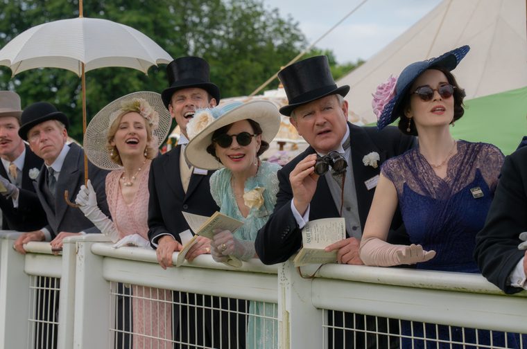 A group of toffs dressed in 1930s suits and dresses stand by a fence, watching a horse race. One man in a top hat is holding binoculars and one woman in a pink dress is holding an umbrella.
