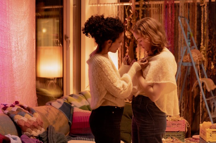A smiling mixed race woman and a white woman stand next to each other, each of them looking down and holding each other's white tops. They are in a cluttered and colourful room full of lamps and knitwear.