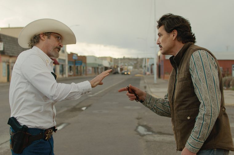 On the left, a man in his late forties wearing glasses and a cowboy hat holds his right hand up to placate a similarly aged man opposite him. The other man is pointing at him and is wearing a brown gilet. They are standing in an empty road of a small dilapidated American town.