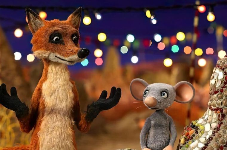 A stop motion animated fox holds out his hands as he talks to a grey, stop motion animated mouse. Fairy lights hang in the background under a dark blue sky.