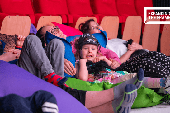 Young girl smiles at camera next to other children and adults laying on bean bags in a cinema, watching a film
