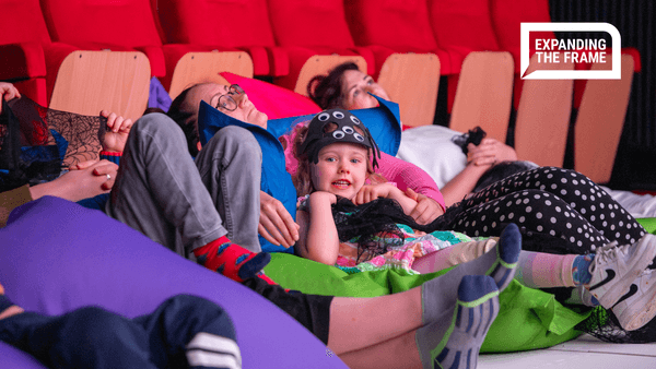 Young girl smiles at camera next to other children and adults laying on bean bags in a cinema, watching a film