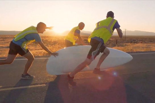 Three people in high-visibility safety vests working together to move what appears to be a large white cylindrical object, possibly a wind turbine blade component, along a road at sunset or sunrise. The scene is set in what looks like a rural or desert area with rolling hills in the background and dramatic golden lighting.