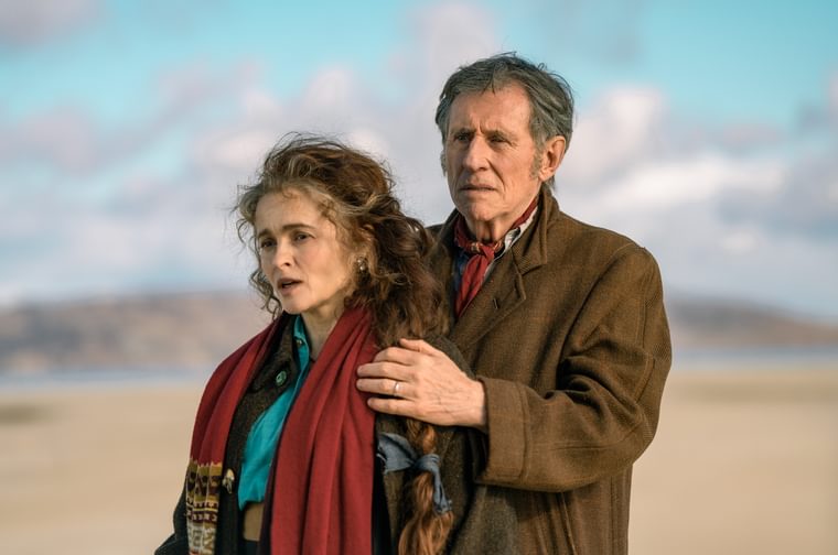An man in his seventies wearing a long brown coat holds the shoulders of a woman in here sixties, who is wearing a red scarf. They are both standing on a beach and staruing off to the left of frame.