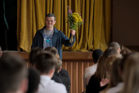 A man is holding what appears to be a bouquet of yellow flowers (possibly daffodils or similar spring flowers with some purple accents). They're wearing a dark jacket over what looks like a graphic t-shirt, and they appear to be smiling while addressing an audience seated in front of the stage. The setting looks like it could be a school auditorium or community center, with golden curtains in the background and wooden paneling visible.