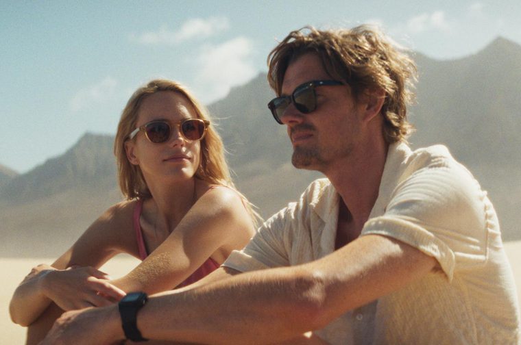 A man and woman in their late thirties sit on a sunny beach, the man staring out to sea as the woman looks at him. They both wear sunglasses.