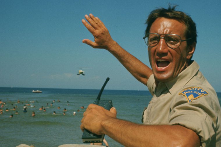 A man in his forties wearing glasses and holding a walkie talkie looks just off screen, shouting to someone. He holds his right hand up in the air, gesturing to a group of swimmers in the ocean behind him.