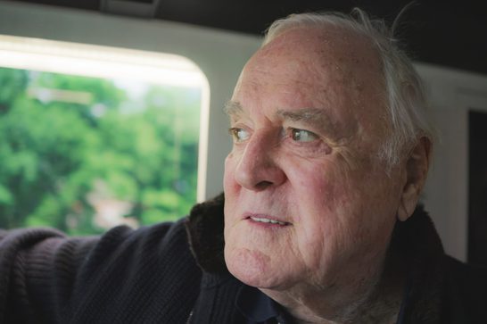 Image of an edlerly man (John Cleese) as he looks off to the left of the image, a window behind him that looks out onto green trees. He is wearing a darkly coloured woolen cardigan.