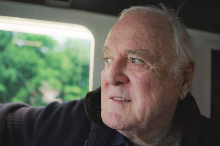Image of an edlerly man (John Cleese) as he looks off to the left of the image, a window behind him that looks out onto green trees. He is wearing a darkly coloured woolen cardigan.