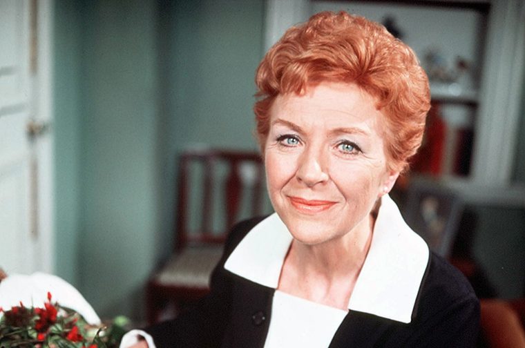 Photo of a woman in her fifties with short auburn hair looking directly into the camera and smiling. She is wearing a black dress with a large white collar and to her right is a bunch of red flowers.