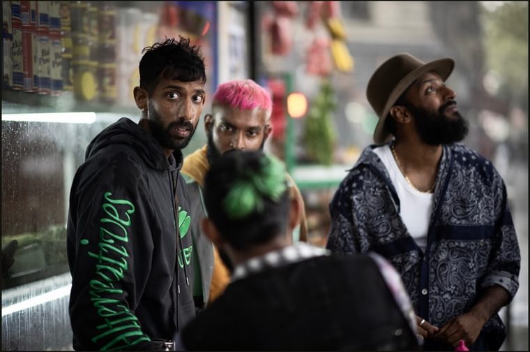 A South Asian man dressed in a black and green hoodie startes off camera. He is surrounded by three other men, one of whom is wearing a trilby hat and staring into the sky.