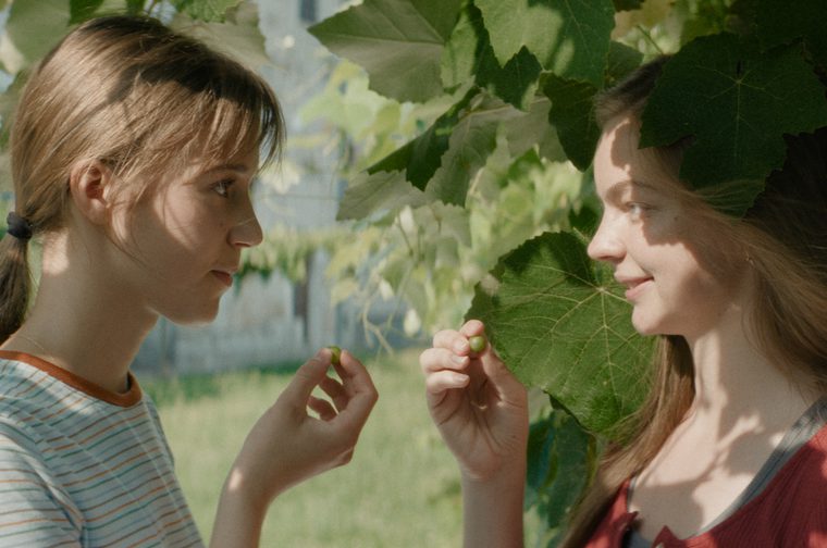 Two young women stand under some leafy foliage, staring into each others' eyes with a half smile on their faces. They are each holding a berry that they've plucked off the trees.