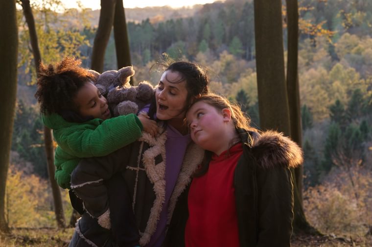 A younger woman holds her two children by her side - one of them is in her arms and clutching a teddy bear, and the other is standing next to her with her head on her shoulder.