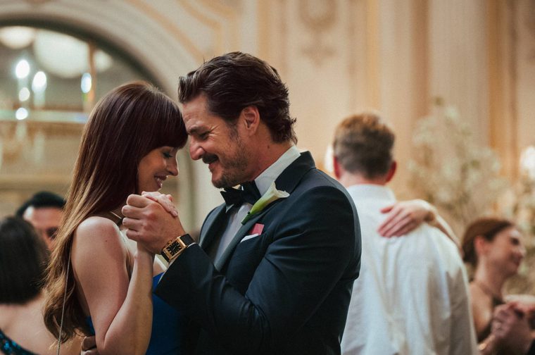 An older bearder man wearing a tuxedo and bow tie slow dances with a younger woman with long brown hair. They are in a posh hall surrounded by other people dancing.
