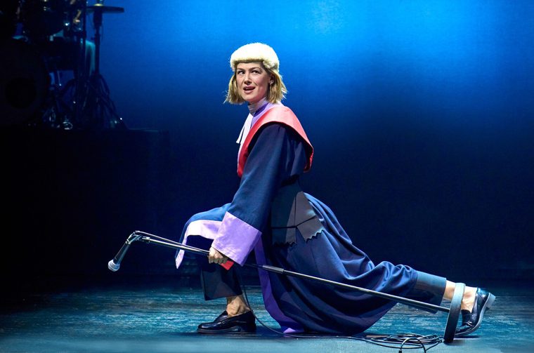 A woman in her late forties wearing a judge's gown and wig kneels down in a rock star pose, holding a microphone stand in her left hand.
