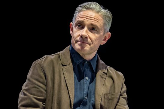 A man in his fifties (Martin Freeman) with grey hair looks off to the left of the frame. He is wearing a blue denim shirt and a light brown jacket.