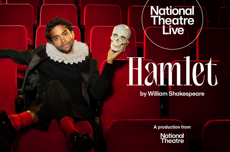 A South Asian man in his thirties sits in an audiotorium with plush red seats. He is wearing a white ruff, a black outfit and red socks, and his feet are crossed over the back of the chair in front of him. He looks directly into the camera. In his left hand he holds up a human skull.