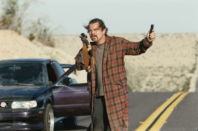 A man in a plaid robe is standing on a desert road next to a dark-colored car. He's holding what looks like a rifle in one hand and a phone in the other hand. The setting appears to be in an arid, desert-like environment with sparse vegetation in the background.
