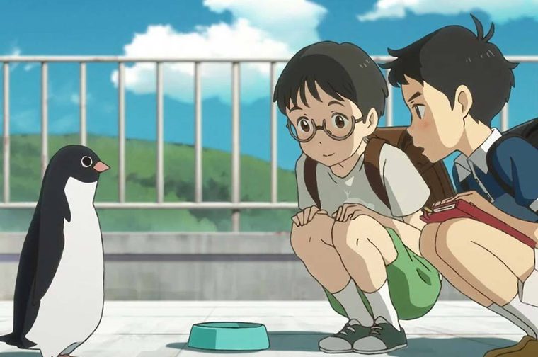 A scene from the animated film 'Penguin Highway' showing two children crouched down observing a penguin on what appears to be a rooftop or terrace. On the left is a young boy with dark hair wearing round glasses, a light-coloured shirt, and green shorts. Next to him is another child with dark hair in a blue jacket, holding what looks like a red notebook or book. They are both looking at a black and white penguin standing near a teal-coloured bowl. The setting features a metal railing in the background with a bright blue sky filled with white clouds and green hills visible in the distance.