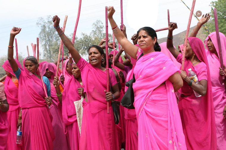A group of Indian woman in bright pink saris stand defiant, each of them raising one hand in the air.