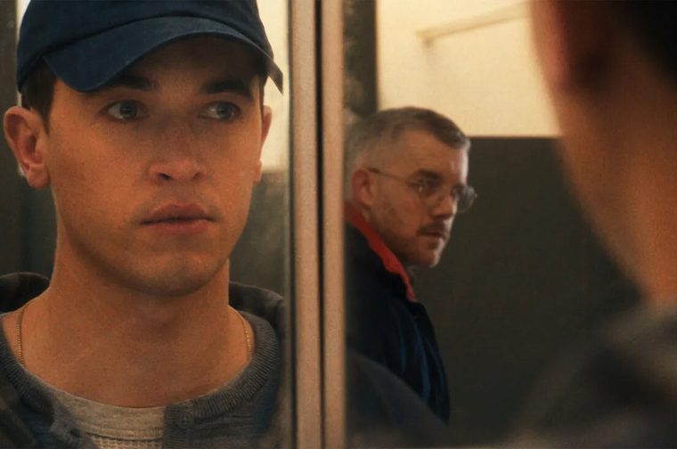 We see two men - one younger man in the foreground wearing a baseball cap and hoodie, and an older man in the background. They stare at each other through a mirror.