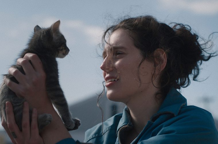 A woman in her twenties with a brunette ponytail and wearing a blue fleece holda a cat in both of her hands, looking directly into its cute fluffy face.
