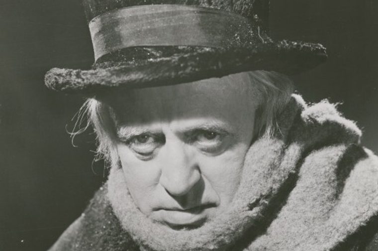 Black and white image of Aklistair Sim as Scrooge, wearing a black top hat, a woolen scarf and a thick dark coat as he stares directly into the camera, a scowl on his face.