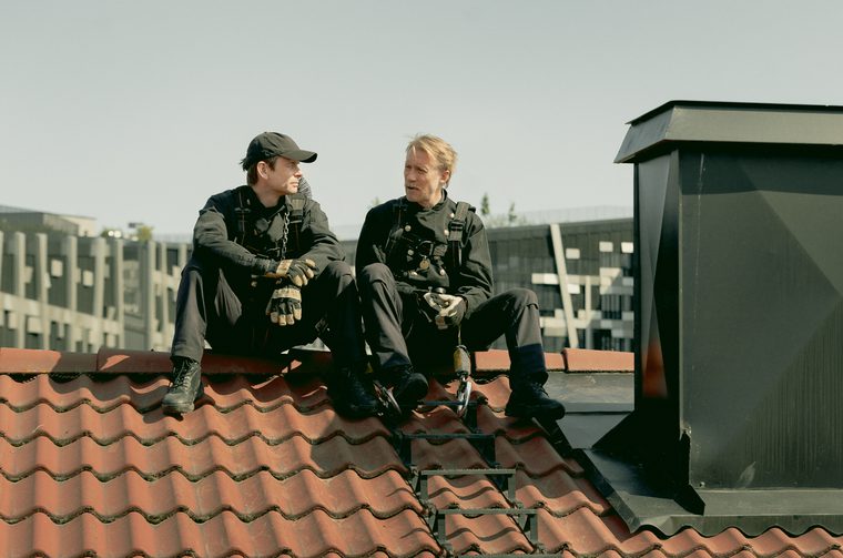 Two men in their forties sit on the top of a roof, next to a large chimney. They are both wearing black chimney sweep outfits and the one on the left is wearing a black cap.