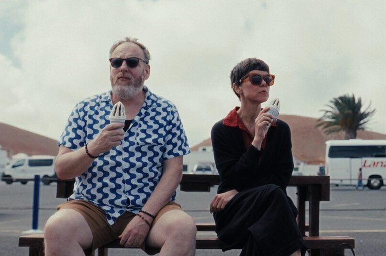 Two people sitting on a bench eating ice cream or popsicles. There's a man in a blue patterned shirt and shorts, and a woman in dark clothing, both wearing sunglasses. They're in what looks like a parking area with buses or RVs in the background, giving it a travel or vacation setting.