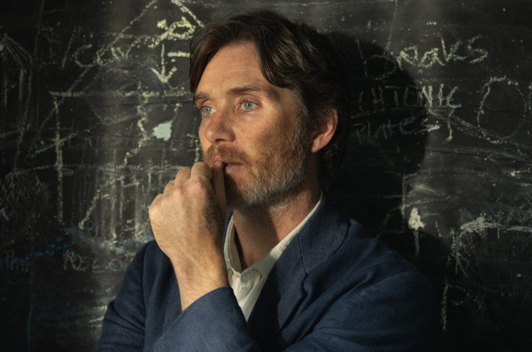 A man in his late forties (Cilliam Murphy) wearing a blue jacket and white shirt with a greying beard stands in ront of a blackboard, staring into the middle distance. His left hand rests on his lips, caught in thought.
