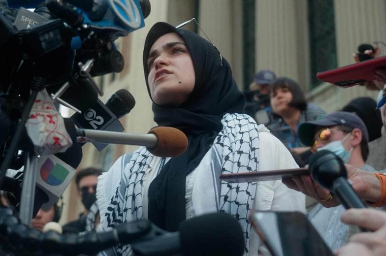 A young Palestinian woman stands in front of a bank and cameras, with journalists surrounding her holding out microphones.