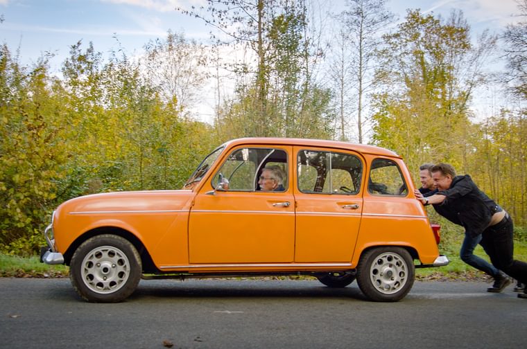 Two fortysomething men dressed in black push an orange mini along a road. Sat in the passenger seat is an elderly man wearing glasses.