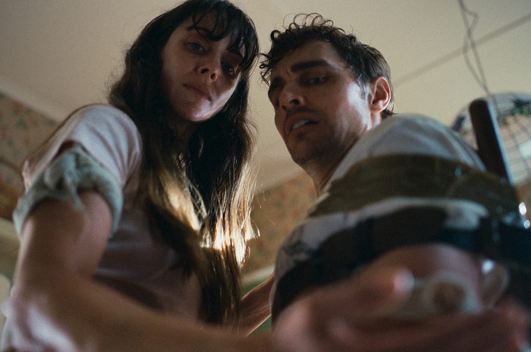 A man and a woman look down at something in the woman's hand that is out of focus. The man is gaffer taped to a chair and they are both sweaty.