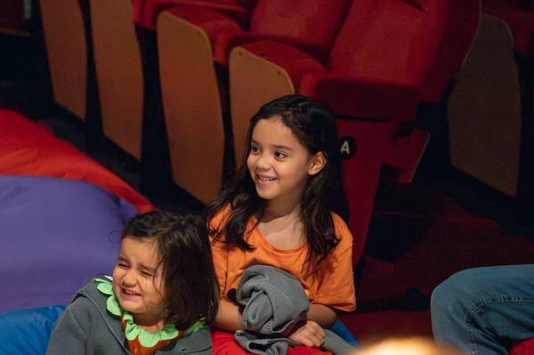 Two little girls sit on beanbags in a cinema smiling to their left, dressed in halloween costumes