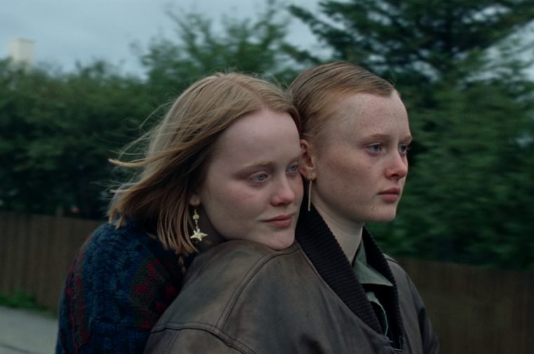 A young woman with long auburn hair rests her chin on the shoulder of another young woman, who has shorter hair and wears a brown leather jacket. They both look off camera, gazing at something in the distance. We see a fence and green hedges behind them.