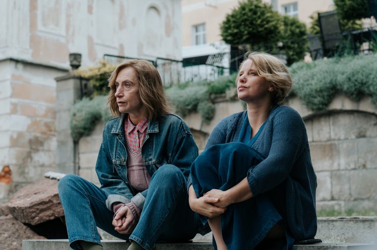 A trans woman dressed in a denim jacket and jeans sits on a step next to a young er blonde haired woman, who is also wearing entirely blue clothing and clutching her knees to her chest. They both look off to the left of the frame.
