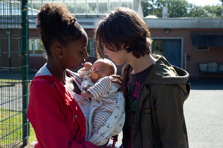A young Black woman in a red hoodie holds a small baby with a dummy in its mouth. A young white boy wearing a dark green hoodie stands next to them. They are both looking down at the baby. On the left of the image is a green fence.
