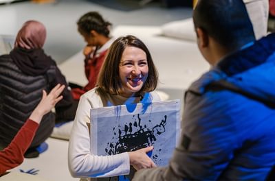 Woman kneeling on the floor holding a piece of paper with a ink explosion on it and showing it to a young boy.