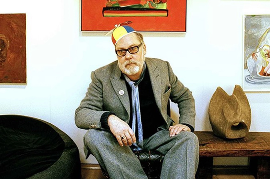 A white man with a beard and glasses (Jim Moir) sits on a table earwing a brightly colour hat a with a propellor on top. He leans forwards with his arms resting on his knees. Behind him are 3 paintings on the wall.