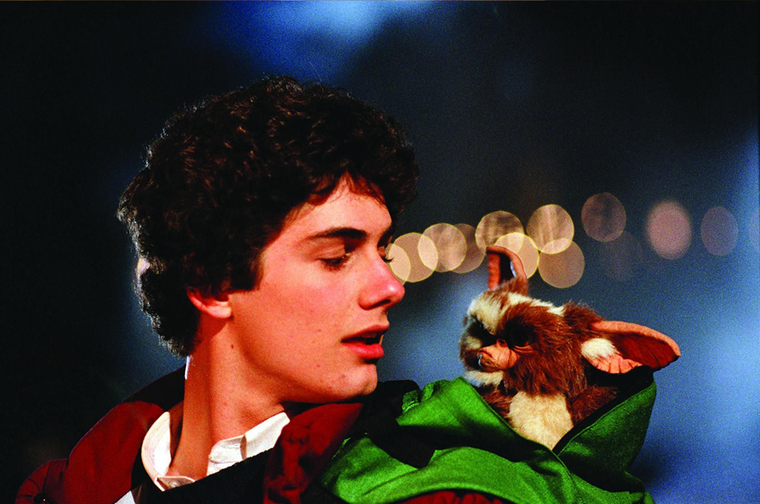 A young white man with dark hair looks at a furry little creature with big ears who is appearing out of his open backpack.