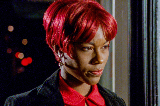 A black woman with shiny red hair and wearing earrings looks to the right - she's wearing a black jacket over a red shirt.