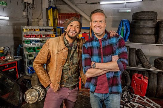 Two men smiling in a garage workshop surrounded by car parts, tools, and stacked tires. The man on the left wears a tan leather jacket and beanie, while the man on the right wears a blue and burgundy plaid shirt with his arms crossed.