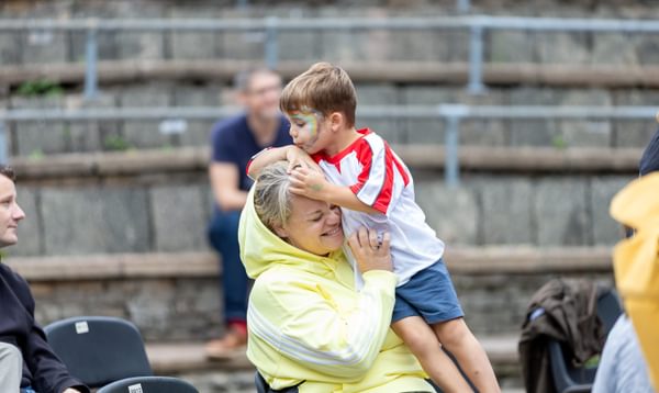 Mum and her son smiling outside. The son is kissing the top of his mum's head and the mum laughs