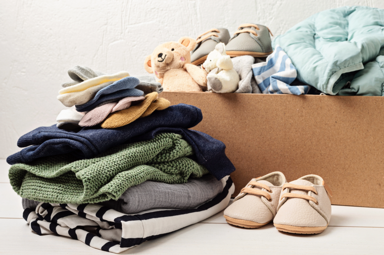 A cardboard box filled with baby items including folded clothes, stuffed animals, shoes, and blankets, with additional baby shoes placed beside the box