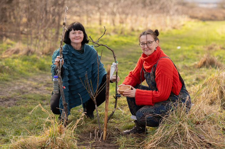 Two women kneeling down in a park. The woman on the right is wearing a bright red jumper and dungarees. The woman on the left is earing a blue poncho and black trousers. They are both holding big sticks which have recycled plastic bottles tied to them.